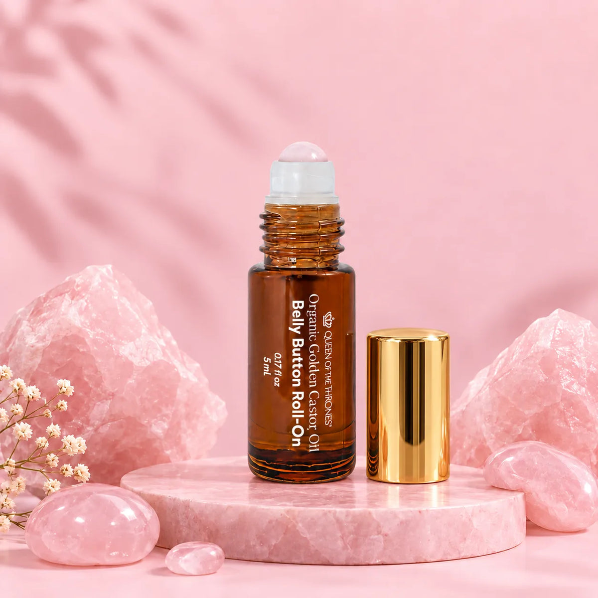 5mL Belly Button Castor Oil Mini Rose Quartz Roll-On on a pink pedestal with its gold cap beside it